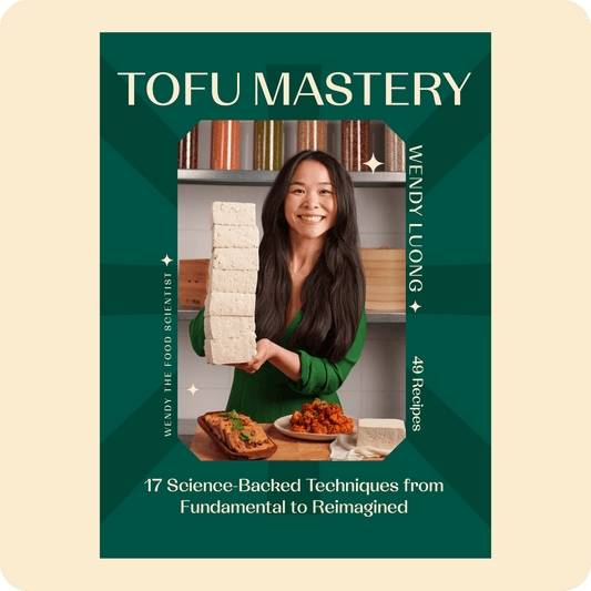 Book cover of 'Tofu Mastery' by Wendy Luong with a woman holding a stack of tofu and tofu dishes on the table in the kitchen.